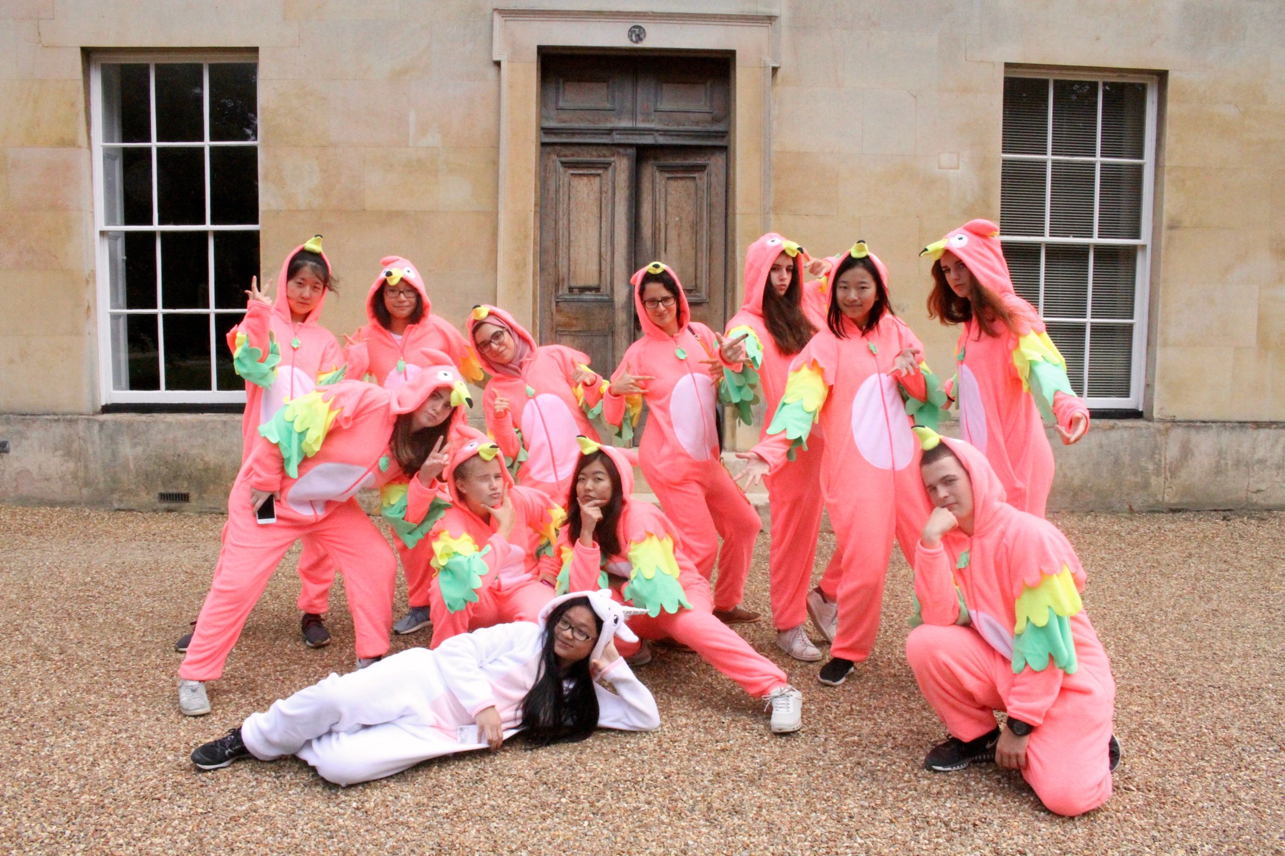 Students at Reach Cambridge pose in bright pink parrot onesies, showcasing creativity and fun in front of a historic building.