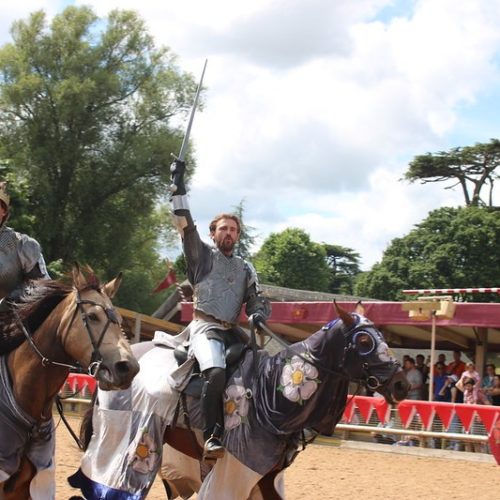 Two knights in armor riding horses and raising their swords during a live historical re-enactment at Warwick Castle, with spectators watching in the background.