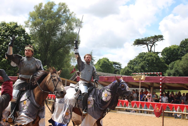 Two knights in armor riding horses and raising their swords during a live historical re-enactment at Warwick Castle, with spectators watching in the background.