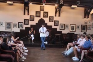 Students participating in a debating session at the Cambridge Union, with a student speaking while others listen attentively in a historic, wood-panelled room adorned with portraits.